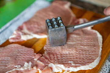 A woman tenderizes fresh raw pork chops with a metal mallet on a wooden cutting board. The process of cooking a homemade meal in the kitchen. The process of cooking schnitzel. 