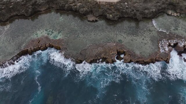 Spectacular top-down aerial drone view of turquoise Pacific waves crashing over a vibrant coral reef in Tonga. A high-contrast scene of white surf and deep blue tropical waters.