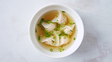 Minimalist overhead shot of Chinese wonton soup. Delicate shrimp and pork dumplings in a clear, golden broth garnished with fresh green onions in a white bowl.