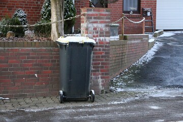 A garbage can, covered with a little snow, stands against the wall on the sidewalk and is ready for collection