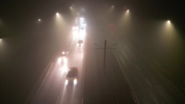 Foggy urban street scene at night features illuminated vehicles navigating through mist, highlighting the atmospheric depth and gradual light changes, camera pans across the roadway