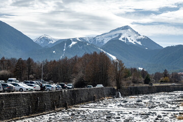 Majestic snow-covered mountains and ski resort rise above a tree line and a rocky riverbed, with parked cars visible on the bank. Bansko, Bulgaria.
