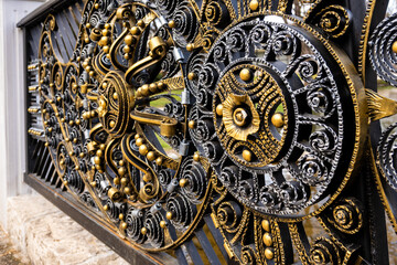 Extreme close-up of a golden and black ornate wrought iron fence, highlighting its intricate three-dimensional decorative swirls, beads, and metallic textures in Razlog, Bulgaria.