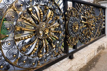 Angled wide shot of ornate golden and black wrought iron fence with intricate patterns, positioned along a stone embankment next to a paved path in Razlog, Bulgaria.