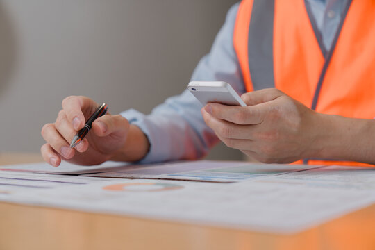 Engineer or construction worker wearing safety vest, holding a smartphone and pen while reviewing documents on a desk. Concept of project management, inspection, and modern workplace technology.