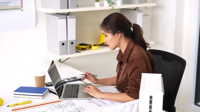 Young female engineer designer or contractor working on drawing plans analyzing structure on laptop computer at construction site office. copy space.
