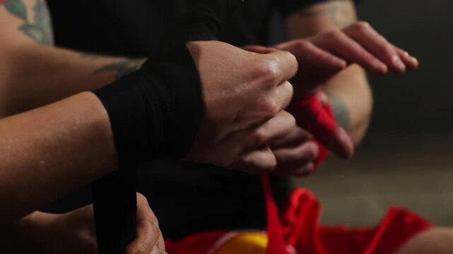 Boxers use special wraps to protect muscles from damage in hall. Partners prepare to begin warming up and practicing skills sitting in spacious boxing gym