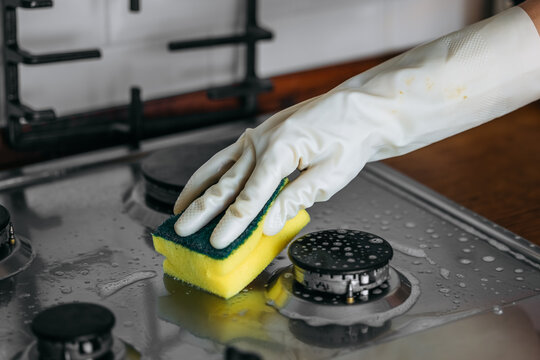 Person wearing rubber gloves is cleaning a stainless steel gas stove with a sponge, showcasing the importance of kitchen hygiene and maintenance in a domestic environment