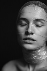 Black and white portrait of a woman with closed eyes, her face and neck wrapped in plastic. A powerful conceptual image exploring themes of restriction, fragility, and environmental impact.