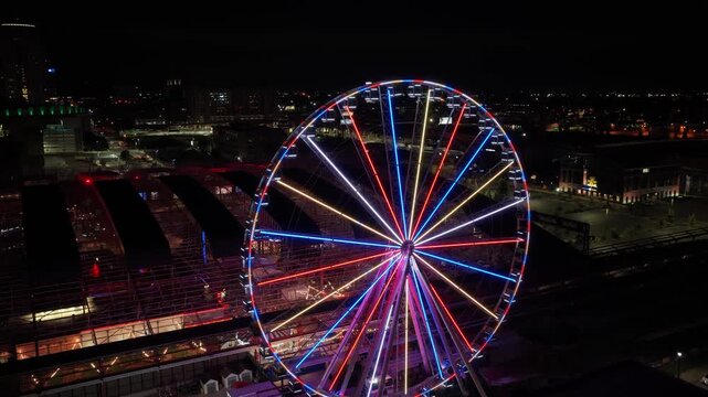 Cinematic nighttime drone orbit of the illuminated Ferris wheel at Union Station in St. Louis, Missouri, with neon lights, city skyline, and the Gateway Arch visible in the background.