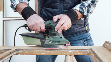 Craftsman. Adult carpenter using an electric sander to smooth an old wooden window. Construction industry, carpentry, housework do it yourself. Restoration.