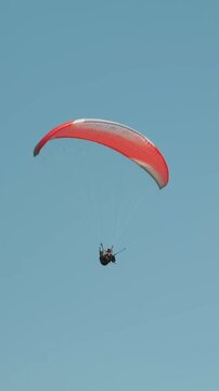 Person gliding on open red parachute against blue clear sky. Vertical video