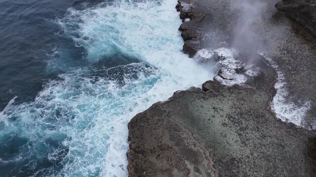 Dramatic top-down aerial drone view of powerful Pacific waves crashing against the rugged volcanic rocky coastline of Tonga, creating beautiful white foam patterns.