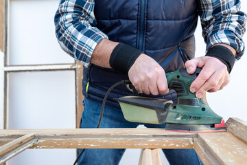 Craftsman. Adult carpenter using an electric sander to smooth an old wooden window. Construction industry, carpentry, housework do it yourself. Restoration.