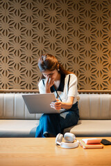 Woman seated on a sofa using a laptop, surrounded by headphones and notebooks, in a stylish room with a geometric patterned wall.