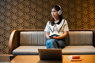 Woman sitting on a sofa with headphones, holding a smartphone, in a modern lounge with a geometric wall and laptop on the table.