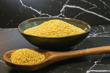 a dark ceramic bowl heaping with raw yellow millet, with a wooden spoon full of grains resting in the foreground on a black marble surface.