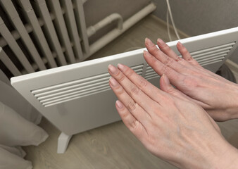 Woman warming hands near convection heater with radiator in background, close up photo