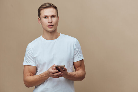young caucasian man holding smartphone wearing white t-shirt standing beige background digital nomad technology communication concept - Powered by Adobe