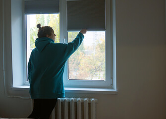 Obraz premium A woman in a sweatshirt raises the blinds on a large window in a house with natural lighting and a view of the street.