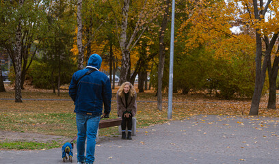 A man in a blue hoodie walks his dog past a laughing woman in a park filled with autumn leaves.