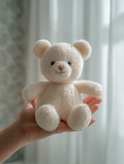 Obraz premium Soft white teddy bear held gently in hand with a bright window and delicate curtain background creating a cozy and warm atmosphere for gift or comfort themes