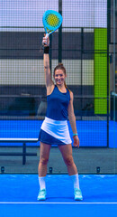 A cheerful, smiling female padel player raises her racket with her arm extended above her head in a gesture of victory and celebration on an indoor blue court.
