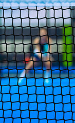 Close-up of a black padel net in focus with a blurred player waiting for a ball in the background on a blue court.
