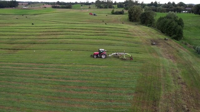 Aerial drone view of a red tractor using a hay rake to shape windrows on a mown grass field, creating striped meadow patterns during summer harvest.