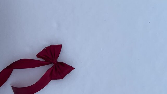 Hand placing a red gift ribbon bow on white snow in the bottom left corner of the frame. Minimal winter background with copy space, festive concept.