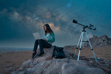 Young woman sitting on rocks using laptop next to a telescope under a starry night sky