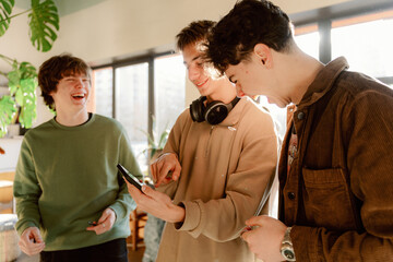Three young friends enjoy a joyful moment together in a bright cafe. They are gathered around a tablet, sharing laughter and ideas while surrounded by greenery.
