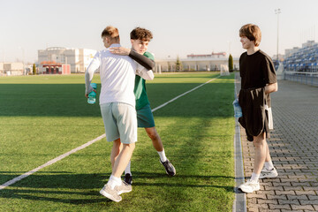 Obraz premium Two young athletes embrace joyfully after a successful soccer practice on a sunny day, while a third friend watches with a smile, standing by the sidelines of the green field.