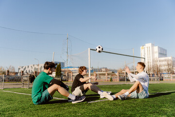 Three friends sit on a lush green soccer field, passing a ball in the bright sunlight. Laughter...
