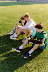 Three young friends sit on a grassy soccer field, catching their breath after an intense game. They...