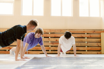 Three young men engage in push-up exercises in a spacious gym with wooden walls. They laugh and motivate each other during their fitness routine, enjoying the energetic atmosphere.