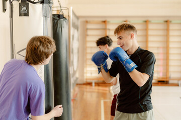 Two young men engage in boxing training in a spacious gym. One practices with a punching bag while...