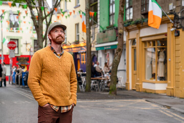 An Irish man in a yellow sweater stands in the heart of Galway's Latin Quarter on Quay Street, a vibrant pedestrian area famous for its colorful storefronts and lively atmosphere.