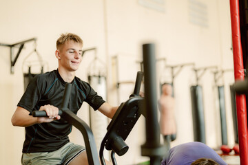 Teenager pedals energetically on an exercise bike in a bustling gym, surrounded by fellow fitness enthusiasts. The space buzzes with motivation and a sense of community.
