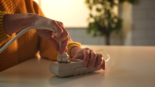 Close up of hands inserting a power plug into a surge protector on a table, with a blurred background.