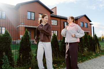 Two young women are jogging along a path next to a lush green fence. They are dressed in comfortable athletic wear, smiling and enjoying a sunny autumn day in a peaceful neighborhood.