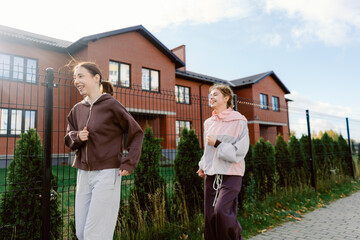 Two friends share laughter while jogging along a peaceful street lined with green bushes. A quaint brick building provides a charming backdrop on this sunny morning.