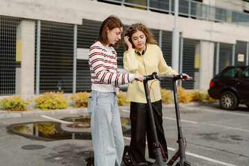Two young women stand in a parking lot, checking their electric scooters. They are excited and...
