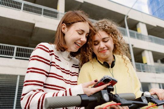 Two young women enjoy a moment together, smiling and focused on their phones. They are standing beside scooters in an urban setting, filled with joy and shared experiences.