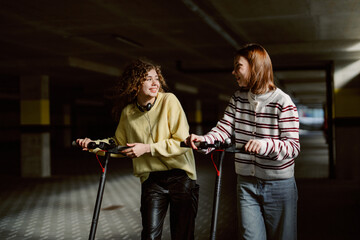 Two young women laugh and chat while riding scooters in a dimly lit parking garage. Their cheerful energy and stylish outfits create a vibrant atmosphere, filled with joy and friendship. © m-art