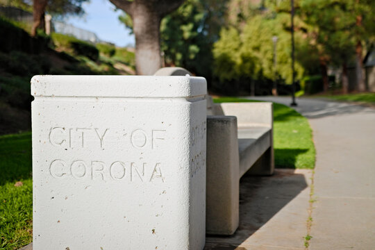 Concrete park bench and waste container marked City of Corona, positioned along a landscaped walkway in a quiet suburban park. - Powered by Adobe