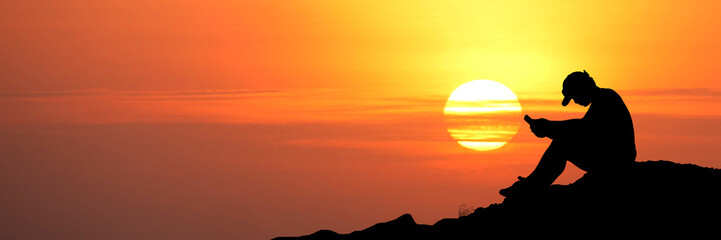 A silhouette of a person using a phone during a warm sunset or sunrise. The sun is a bright ball of fire in the sky, creating a beautiful and dramatic scene. 