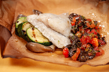 Sea bass fillet baked with zucchini, tomatoes, olives, and herbs served on a plate with parchment paper