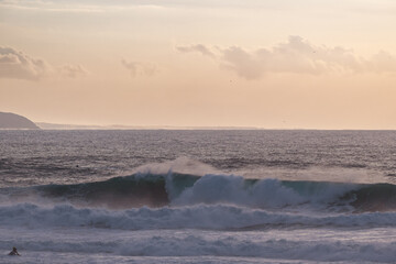 Calm ocean sunset with large waves and distant coastline, surfers nearby