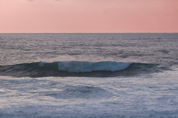 Calm ocean waves roll under a pink sunset sky over the tranquil sea